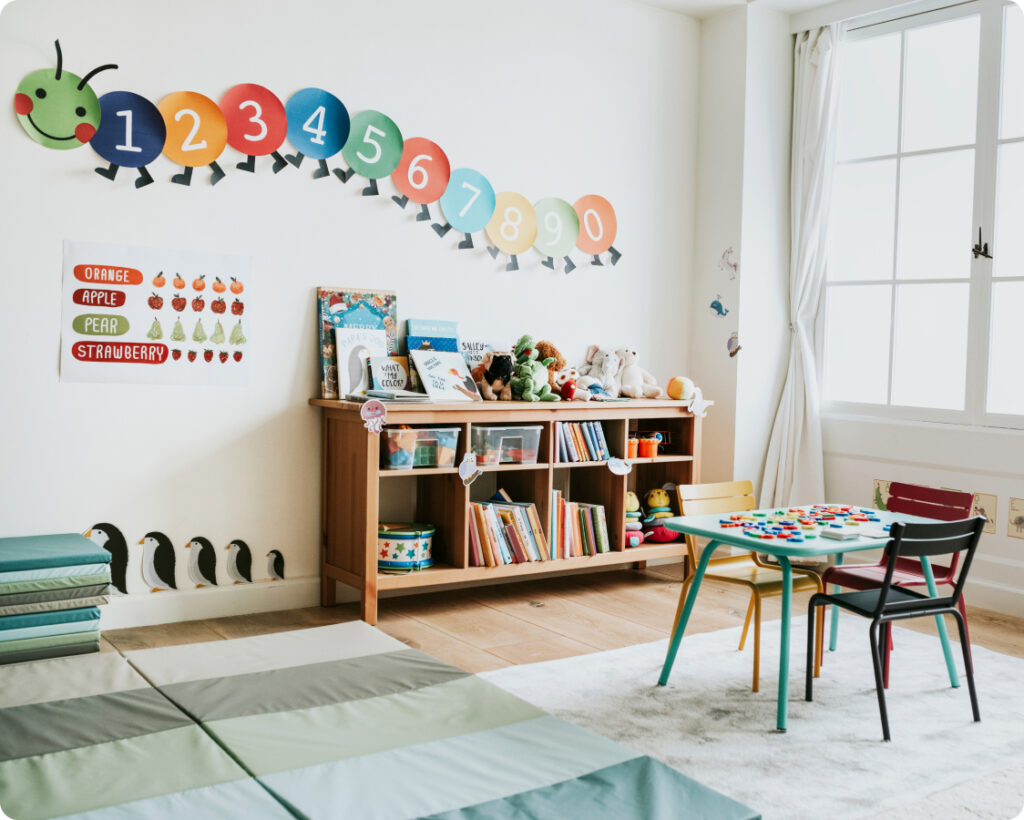 Empty classroom with a play mat, desk with three chairs, and a caterpillar graphic on the wall.